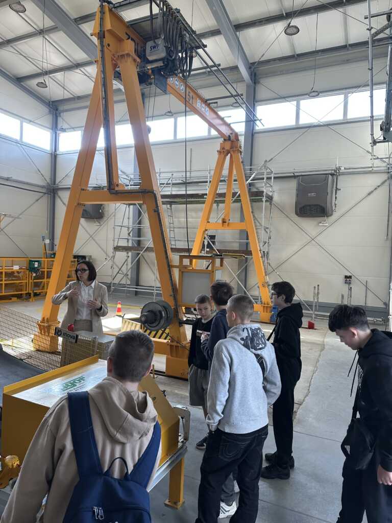 Grupa studentów stoi wokół stołu ekspozycyjnego, słuchając kobiety przemawiającej w warsztacie przemysłowym z dużą żółtą suwnicą w tle.