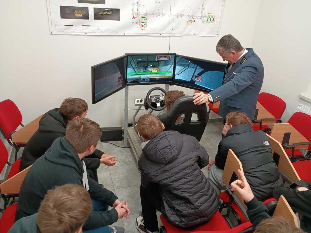 Grupa uczniów siedzących wokół symulatora jazdy z trzema monitorami obserwuje, jak umundurowany instruktor wyjaśnia coś uczniowi korzystającemu z symulatora w klasie.