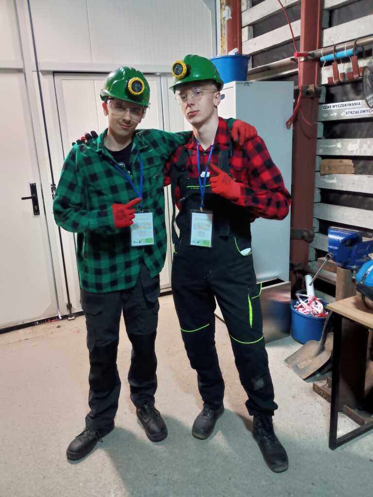 Dwie osoby w zielonych kaskach, okularach ochronnych, koszulach w kratę, czarnych kombinezonach, czerwonych rękawicach i identyfikatorach stoją w pomieszczeniu, ramię w ramię, w warsztacie lub otoczeniu przemysłowym.