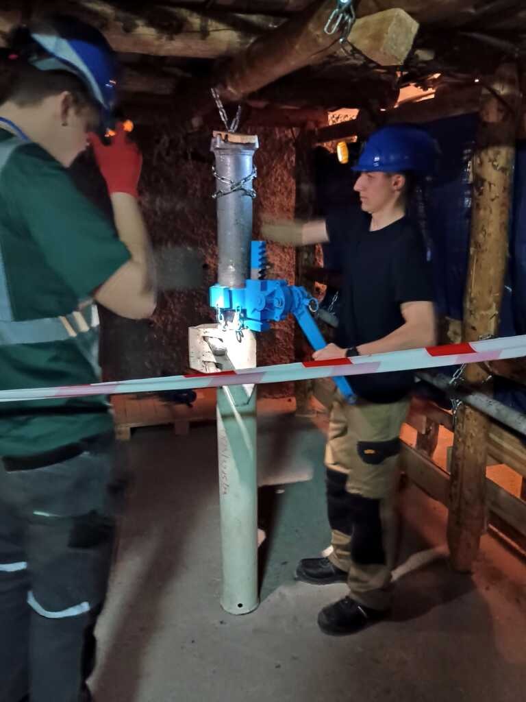 Dwóch pracowników w odzieży ochronnej i niebieskich kaskach obsługuje niebieską obrabiarkę na metalowej rurze w podziemnym, słabo oświetlonym obszarze z drewnianymi podporami; jeden reguluje narzędzie, podczas gdy drugi obserwuje.