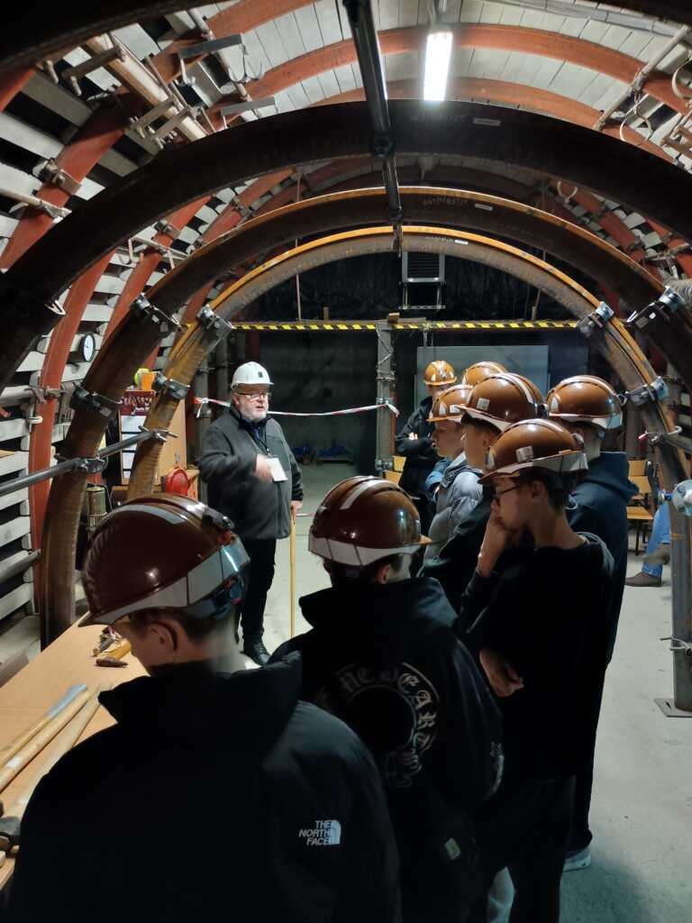 Grupa ludzi w brązowych kaskach i kamizelkach ochronnych słucha przemawiającej osoby na placu budowy tunelu, otoczonej sprzętem i zakrzywionymi metalowymi wspornikami.