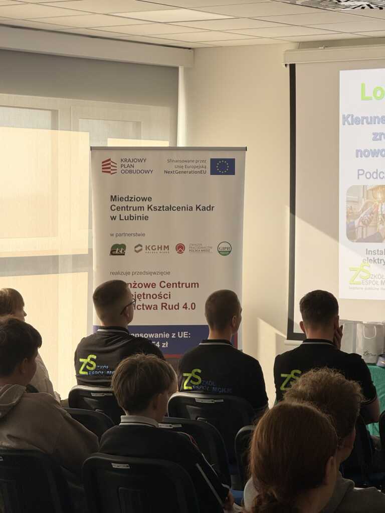 Grupa ludzi siedzi w oświetlonym słońcem pomieszczeniu naprzeciwko ekranu prezentacyjnego i banerów, a niektórzy uczestnicy mają na sobie ciemne koszulki z zielonym logo "zs" na plecach.