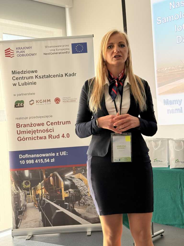 Kobieta o długich blond włosach stoi w pomieszczeniu, przemawiając przed banerem reklamowym na temat szkolenia pracowników w górnictwie. Ma na sobie ciemną marynarkę i plakietkę konferencyjną, a za nią widoczne są slajdy prezentacji i stół.