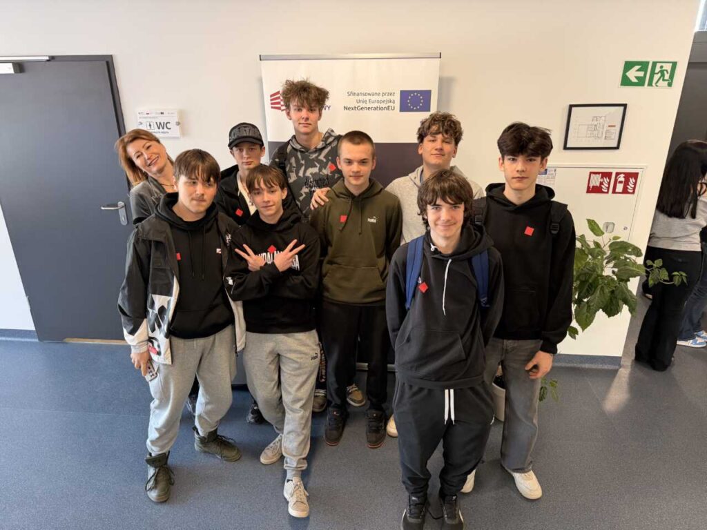 Grupa dziewięciu nastolatków i jednego dorosłego, wszyscy w zwykłych ubraniach, stoją i uśmiechają się w pomieszczeniu przed ścianą z plakatem Unii Europejskiej. Na ścianach widoczne są znaki wyjścia i znaki informacyjne.