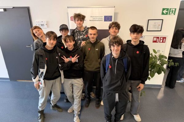 Grupa dziewięciu nastolatków i jednego dorosłego, wszyscy w zwykłych ubraniach, stoją i uśmiechają się w pomieszczeniu przed ścianą z plakatem Unii Europejskiej. Na ścianach widoczne są znaki wyjścia i znaki informacyjne.