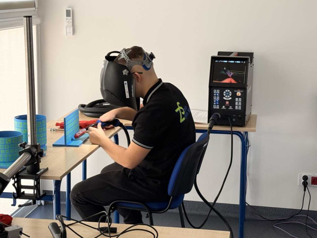 Osoba nosząca gogle VR siedzi przy biurku, trzymając narzędzia spawalnicze i ćwicząc na wirtualnym symulatorze spawania. Ekran na stole wyświetla symulowany proces spawania.