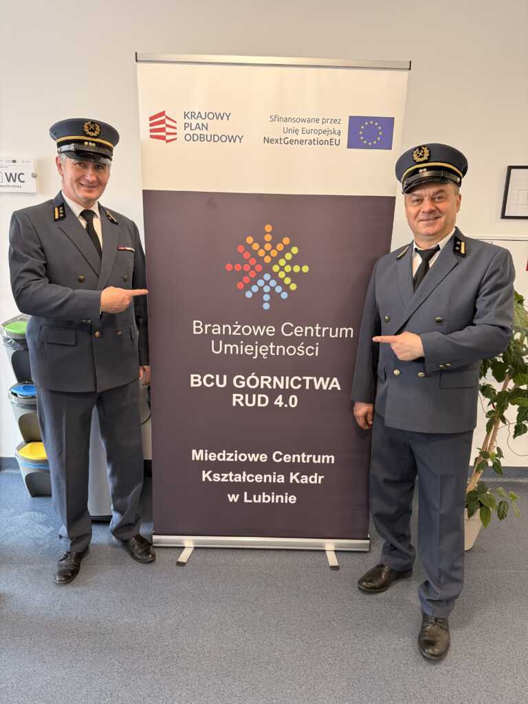 Dwóch mężczyzn w dopasowanych niebieskich mundurach i czapkach stoi po obu stronach znaku, uśmiechając się i wskazując na niego. Znak wyświetla logo i polski tekst o centrum umiejętności w Lubinie, współfinansowanym przez UE.