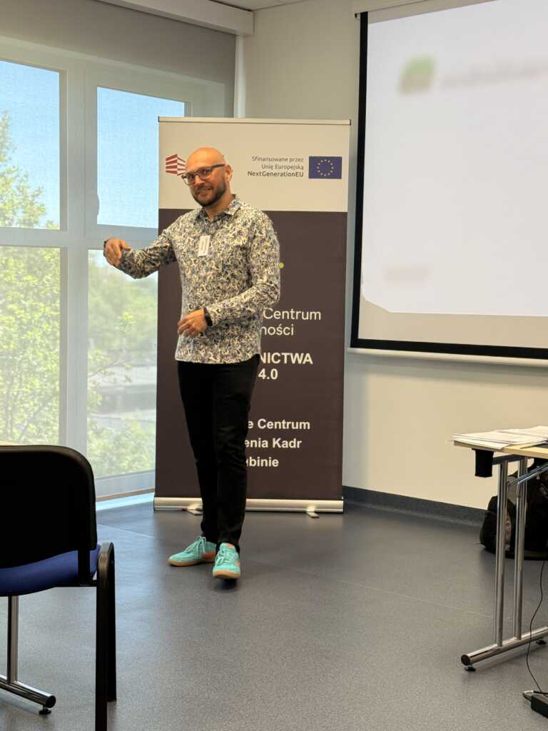Mężczyzna w okularach, z brodą i w turkusowych butach prowadzi prezentację w jasnym pomieszczeniu. Za nim znajduje się baner z logo i ekran projekcyjny. Na pierwszym planie widoczne są krzesła i biurko.